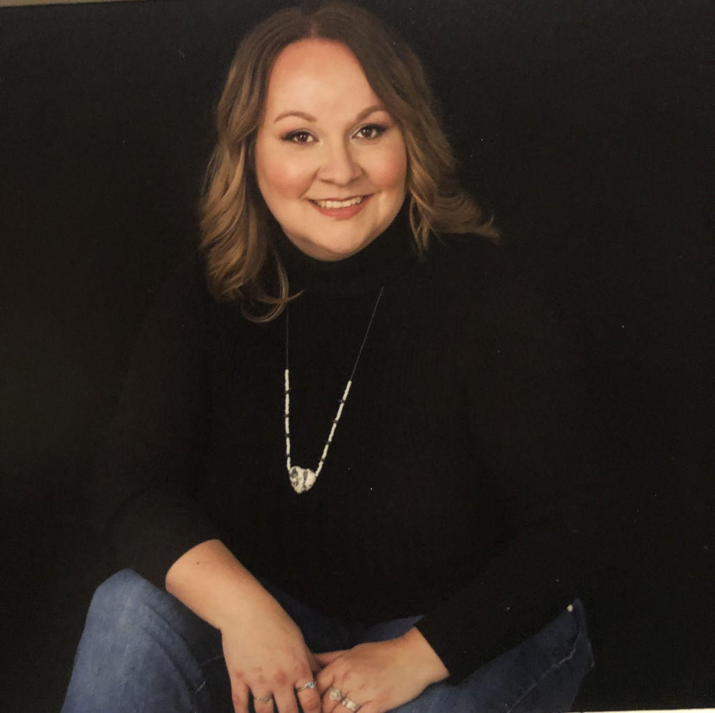 Studio portrait of Amie Arizmendi smiling, wearing a black long-sleeve top against a dark background.