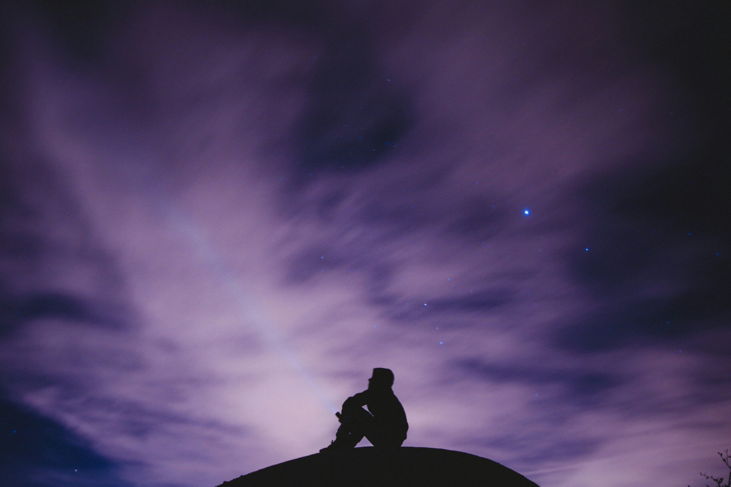 Silhouette of a person sitting on a hill under a dramatic, cloudy purple and dark blue night sky.